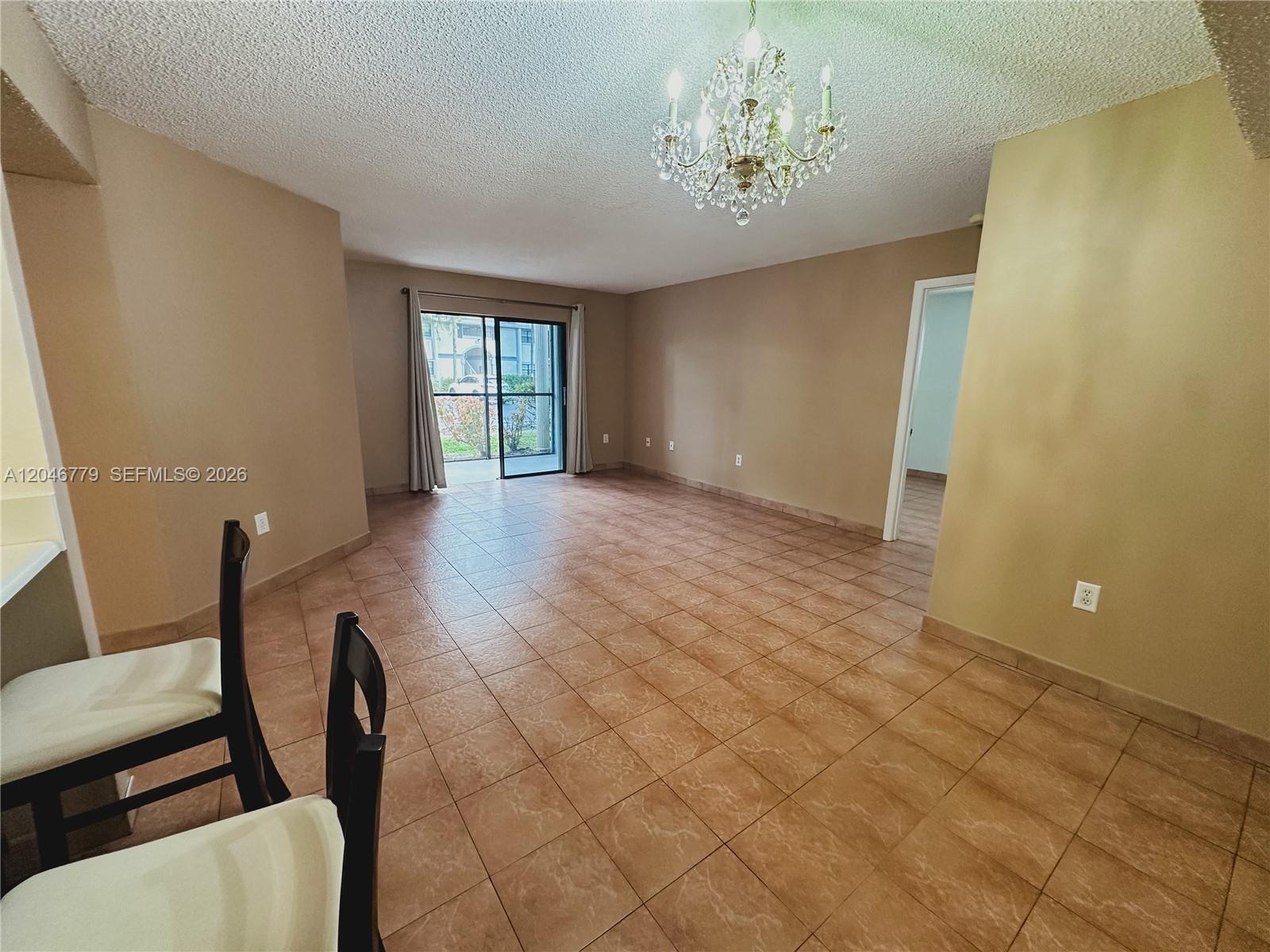 An empty living room features tan walls, tile flooring, a chandelier, and a sliding glass door — 927 NE 199th St # 101, Miami FL 33179, MIAMI, FL