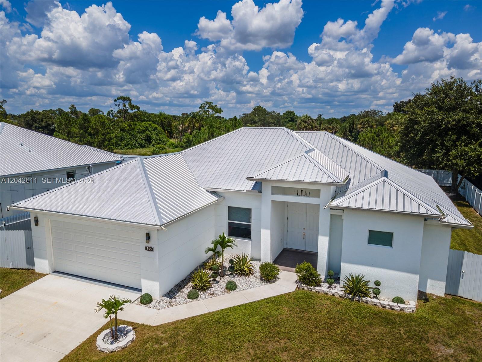 An aerial view shows a white single-story house with a metal roof and landscaping — 365 Ranier Ave, Lehigh Acres FL 33974, LEHIGH ACRES, FL