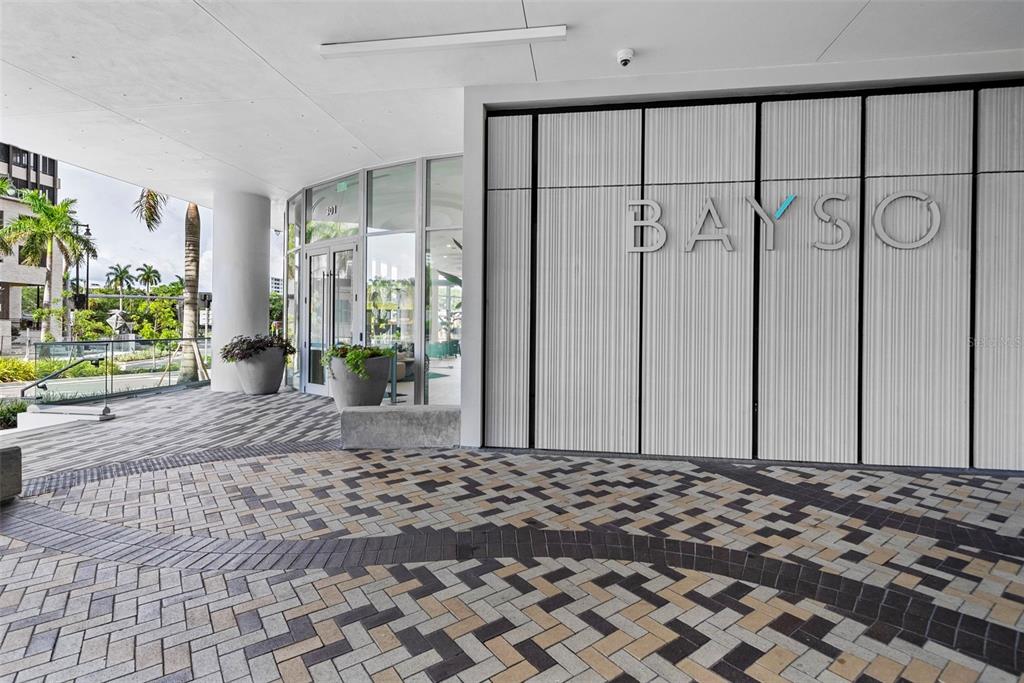The exterior entrance of a modern building features a paved driveway and a sign reading BAYSO — 301 QUAY COMMONS UNIT 1009, SARASOTA, FL