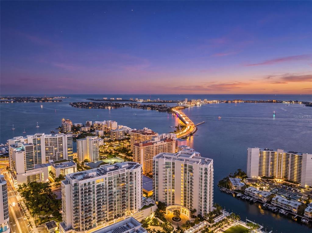 An aerial view shows high-rise buildings and a long bridge spanning across a bay — 301 QUAY COMMONS UNIT 1009, SARASOTA, FL