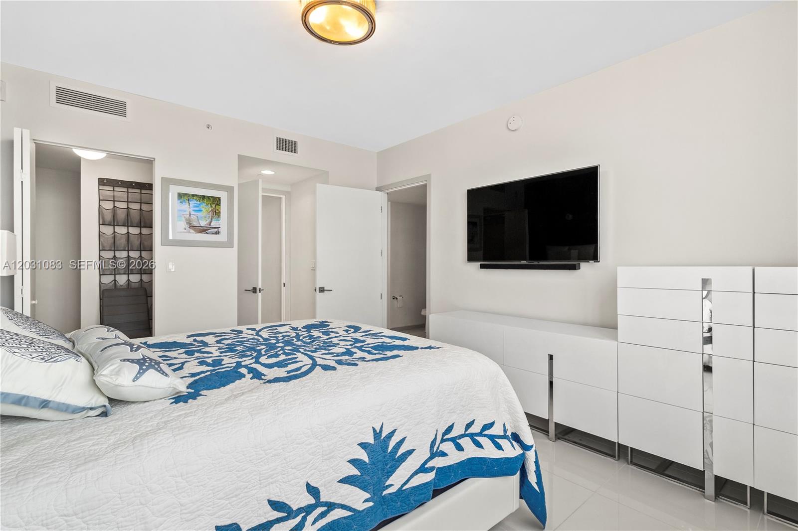 A bedroom features a white bed with a blue patterned quilt and a wall-mounted television — DORAL, FL