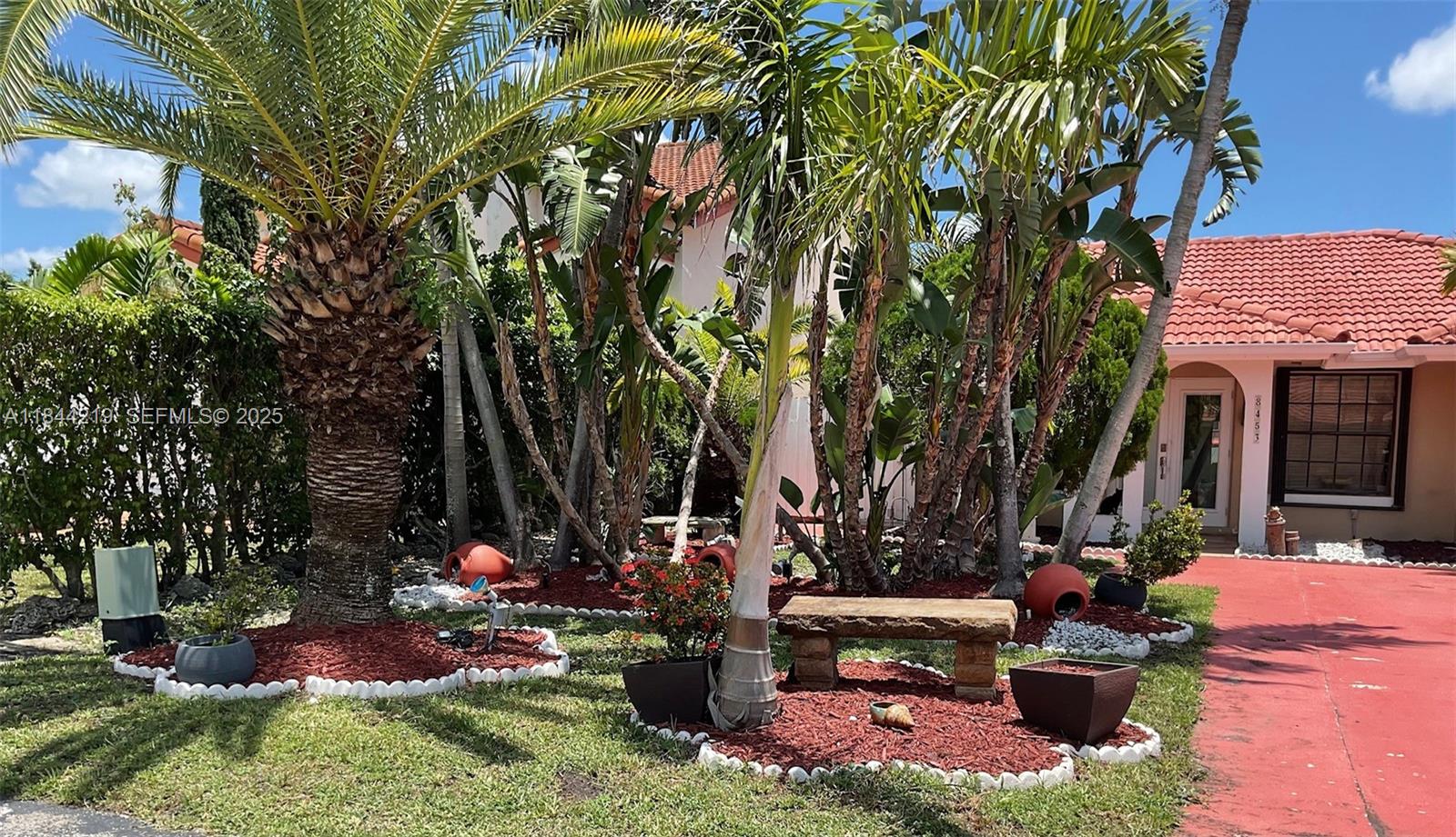 A stone bench sits on a mulched garden bed near palm trees and a paved street — 8453 NW 189 Street Rd, Hialeah FL 33015, HIALEAH, FL
