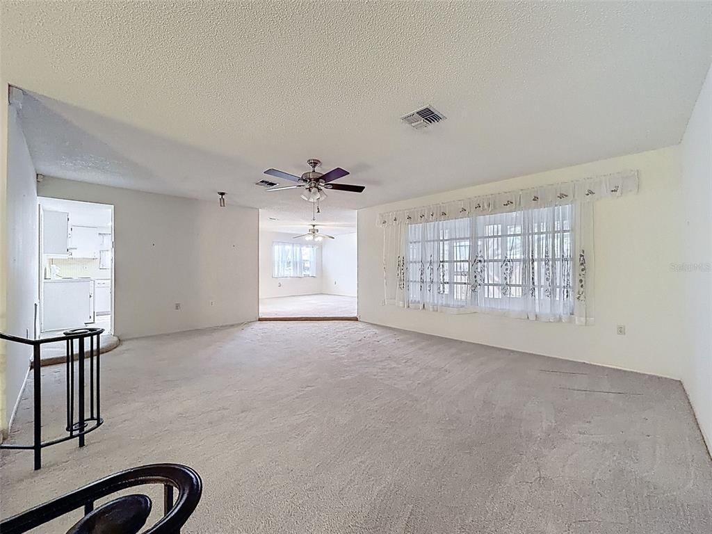 A spacious, carpeted living room features a ceiling fan, large windows with curtains, and white walls — 9811 WOODSTOCK LANE, PORT RICHEY, FL