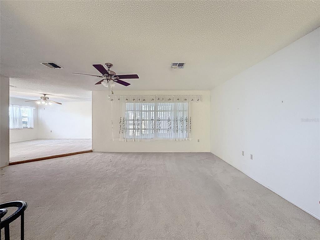 A spacious living room features beige carpeting, a ceiling fan, and a large window with curtains — 9811 WOODSTOCK LANE, PORT RICHEY, FL