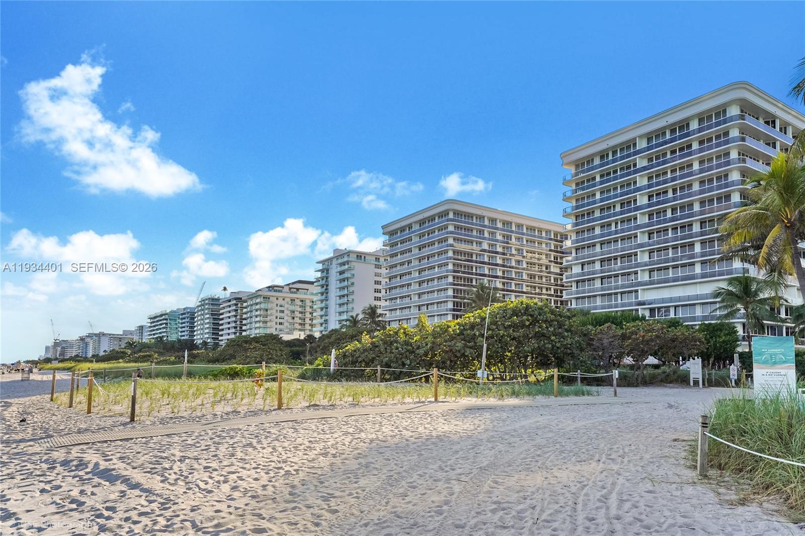 A sandy beach path leads toward a row of high-rise apartment buildings under a blue sky — 9559 Collins Ave # F, Surfside FL 33154, SURFSIDE, FL