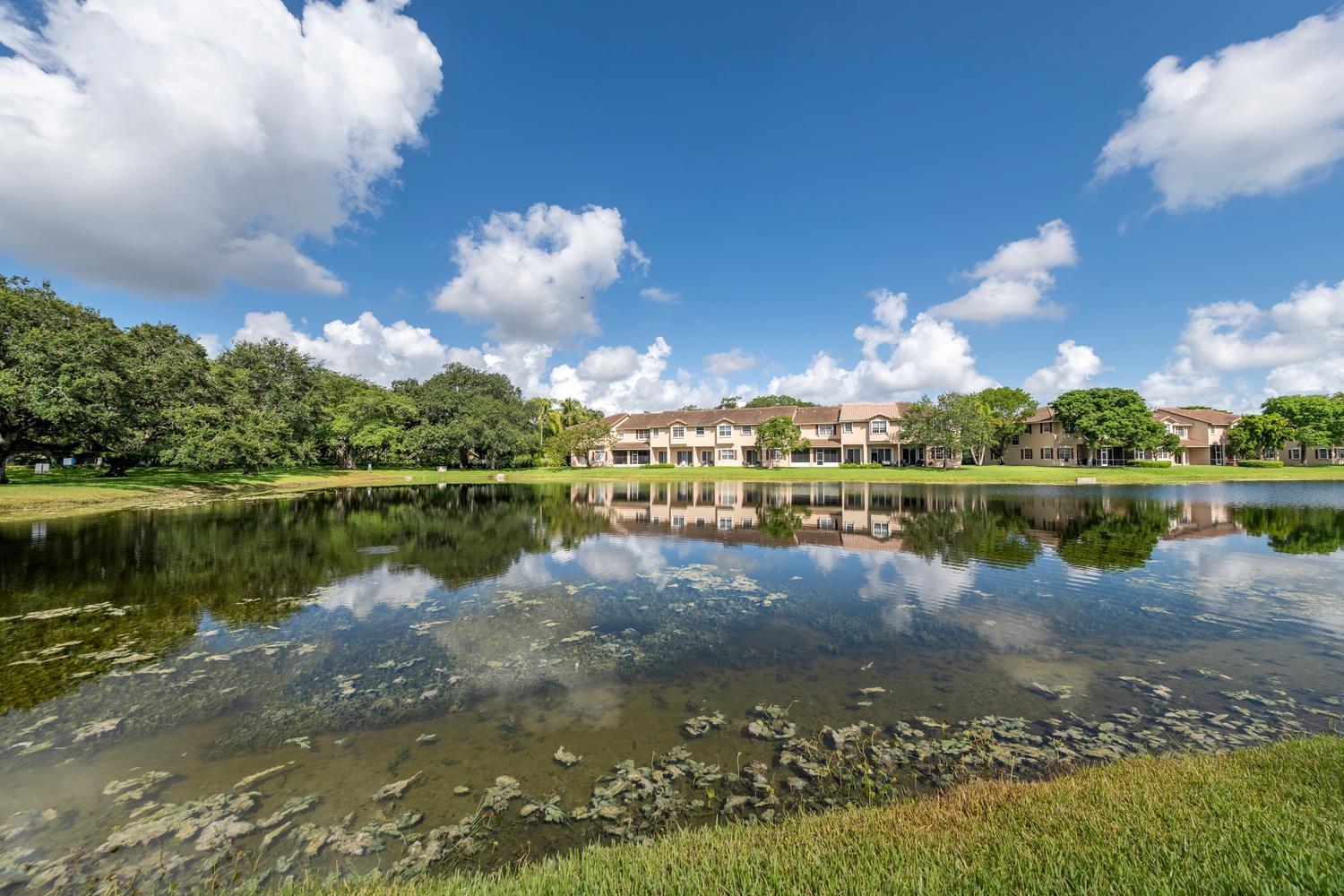 A pond reflects a multi-unit residential building under a blue sky with scattered clouds — 3501 Lakeside Drive, Davie FL 33328, DAVIE, FL