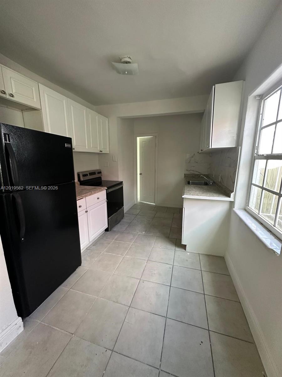 A kitchen features white cabinets, a black refrigerator, a stove, and light-colored tile flooring — MIAMI, FL