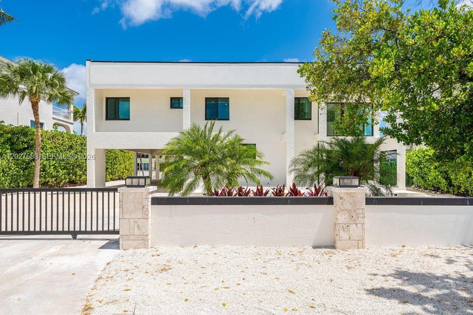 A two-story white modern house features a gated gravel driveway and lush tropical landscaping — KEY LARGO, FL