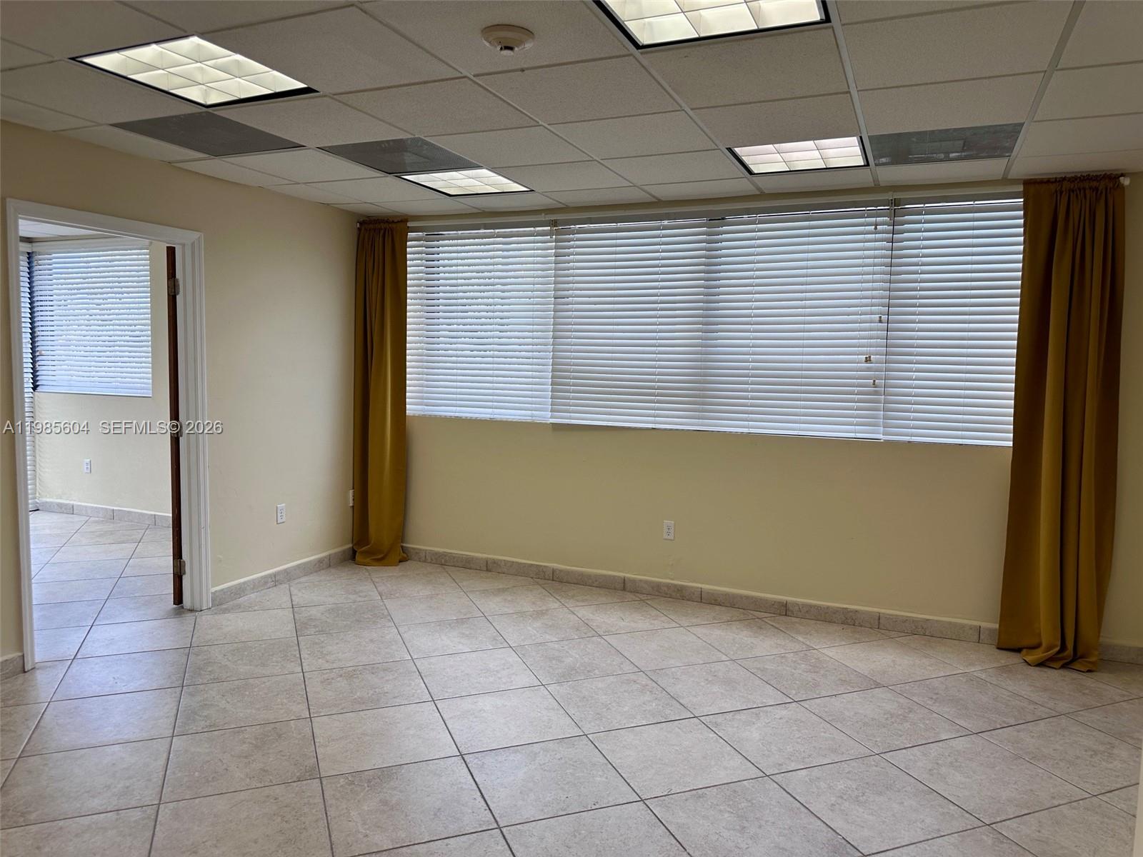 An empty room features beige tile flooring, mustard curtains, and a drop ceiling with lights — 5455 SW 8 ST # 200, Miami FL 33134, MIAMI, FL
