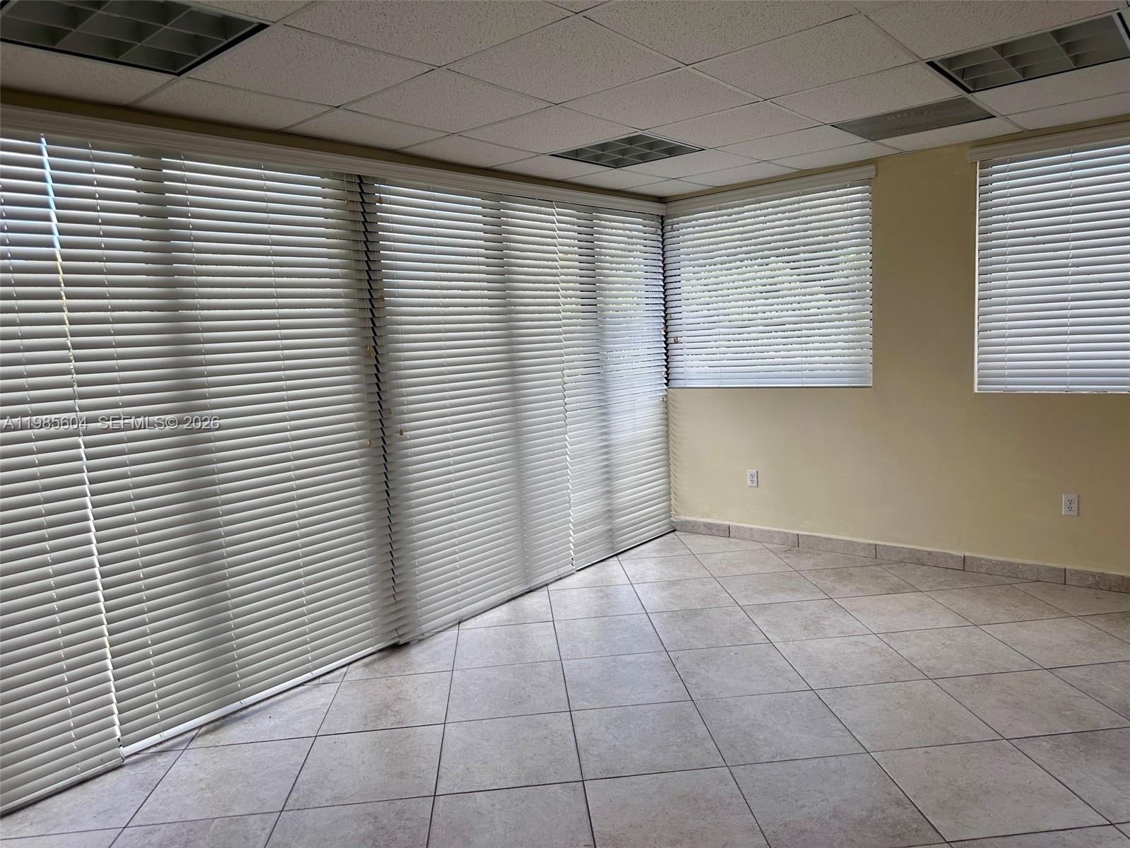 An empty room features tiled flooring, a drop ceiling, and large windows with white blinds — 5455 SW 8 ST # 200, Miami FL 33134, MIAMI, FL