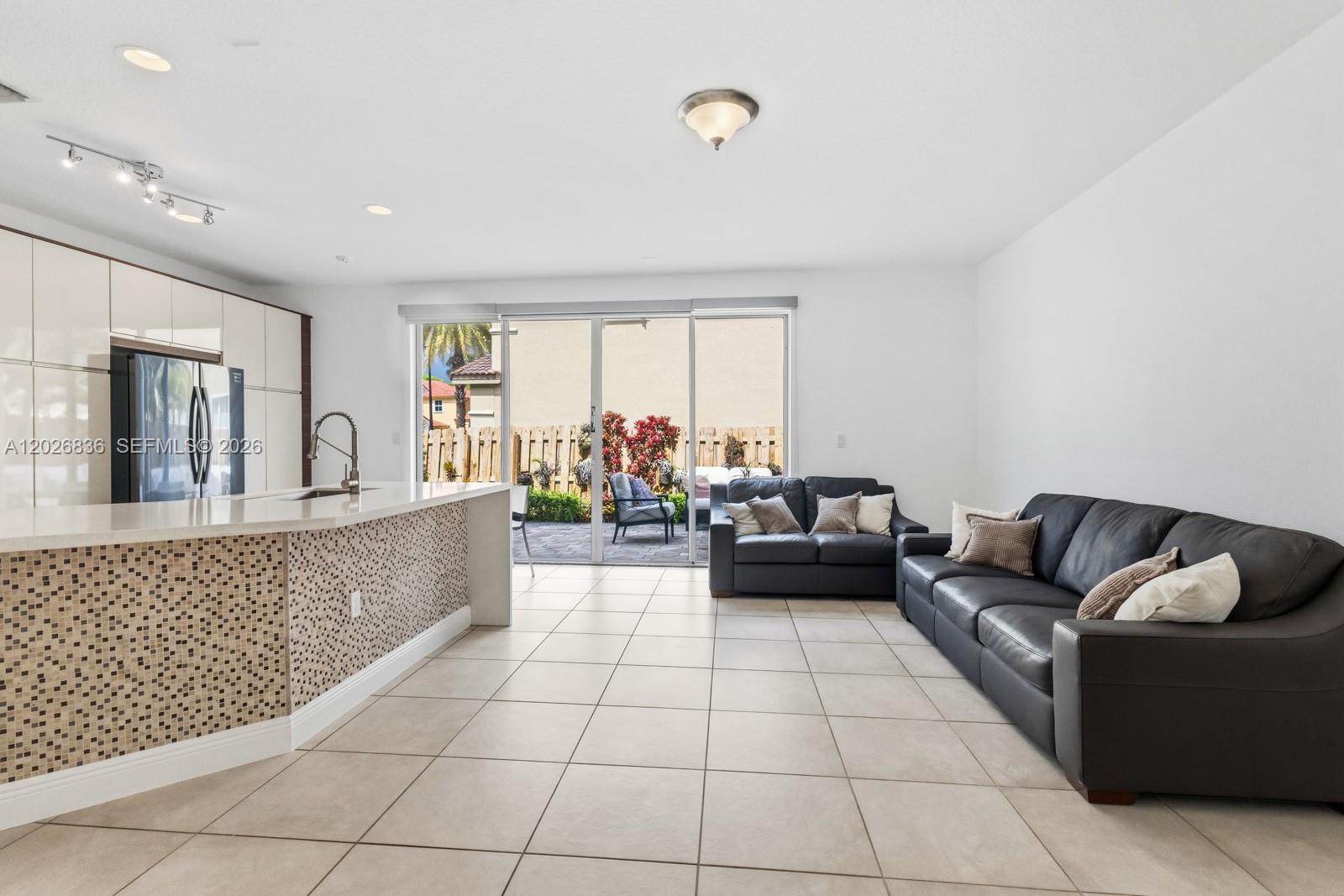 An open-concept kitchen and living area features tile flooring, dark leather sofas, and sliding doors — 8610 NW 110th Ave, Doral FL 33178, DORAL, FL