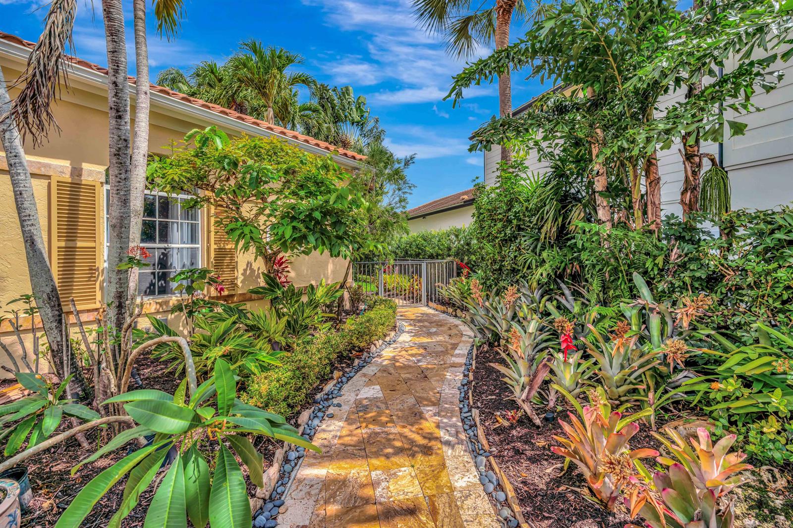 A stone walkway leads past lush tropical landscaping toward a gate between two residential buildings — 186 Golf Village Boulevard, Jupiter FL 33458, JUPITER, FL