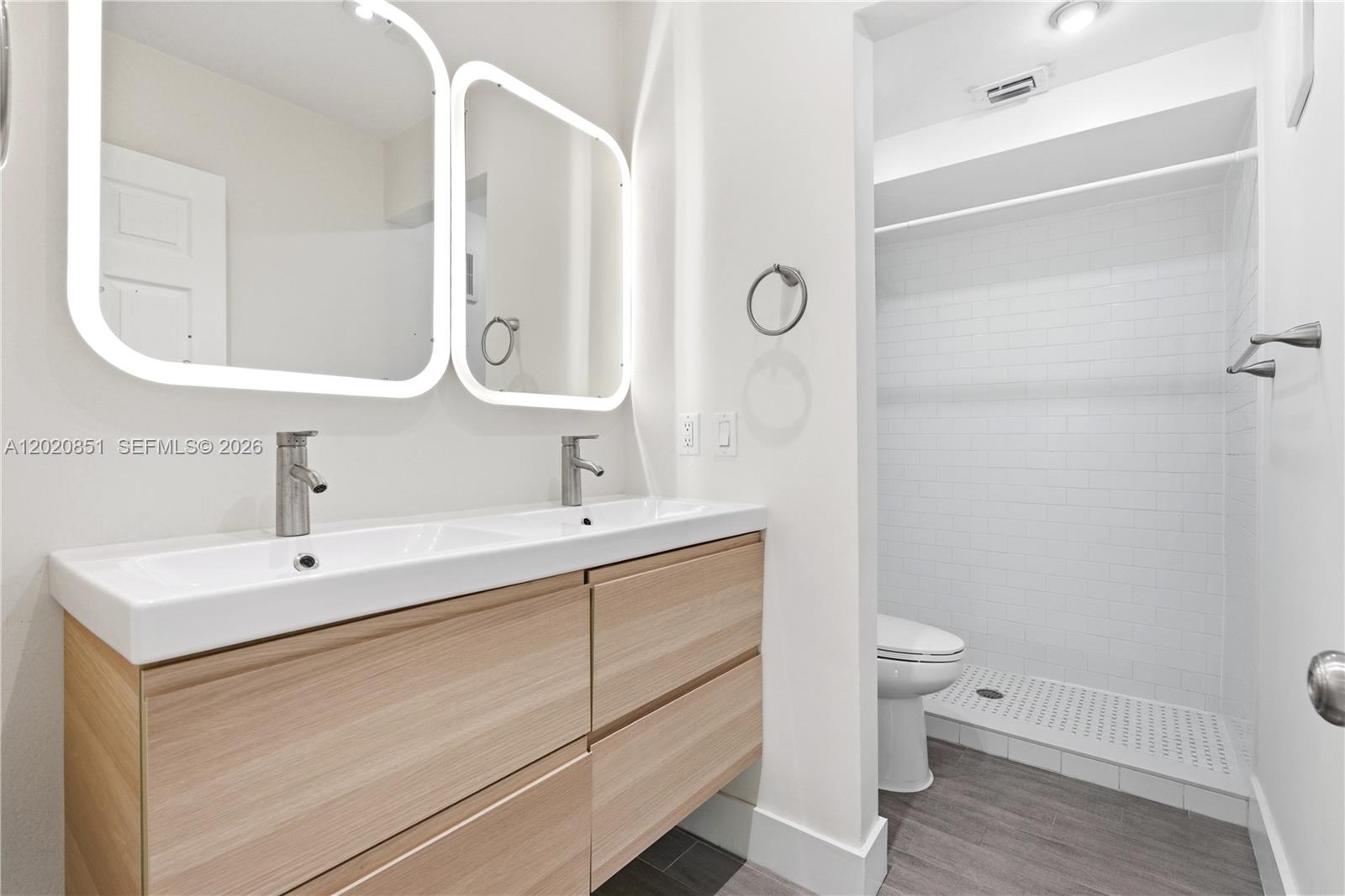 A modern bathroom features a double vanity with light wood cabinets and backlit mirrors — 1200 Mariposa Ave # E104, Coral Gables FL 33146, CORAL GABLES, FL