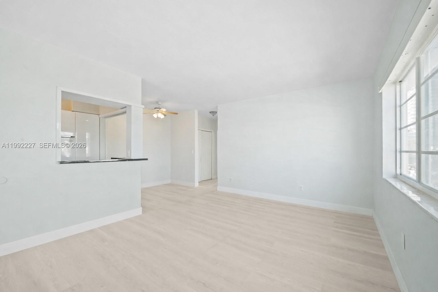 An empty living room features light-colored wood-look flooring, white walls, and a kitchen pass-through window — 7315 Byron Ave # 10, Miami Beach FL 33141, MIAMI BEACH, FL