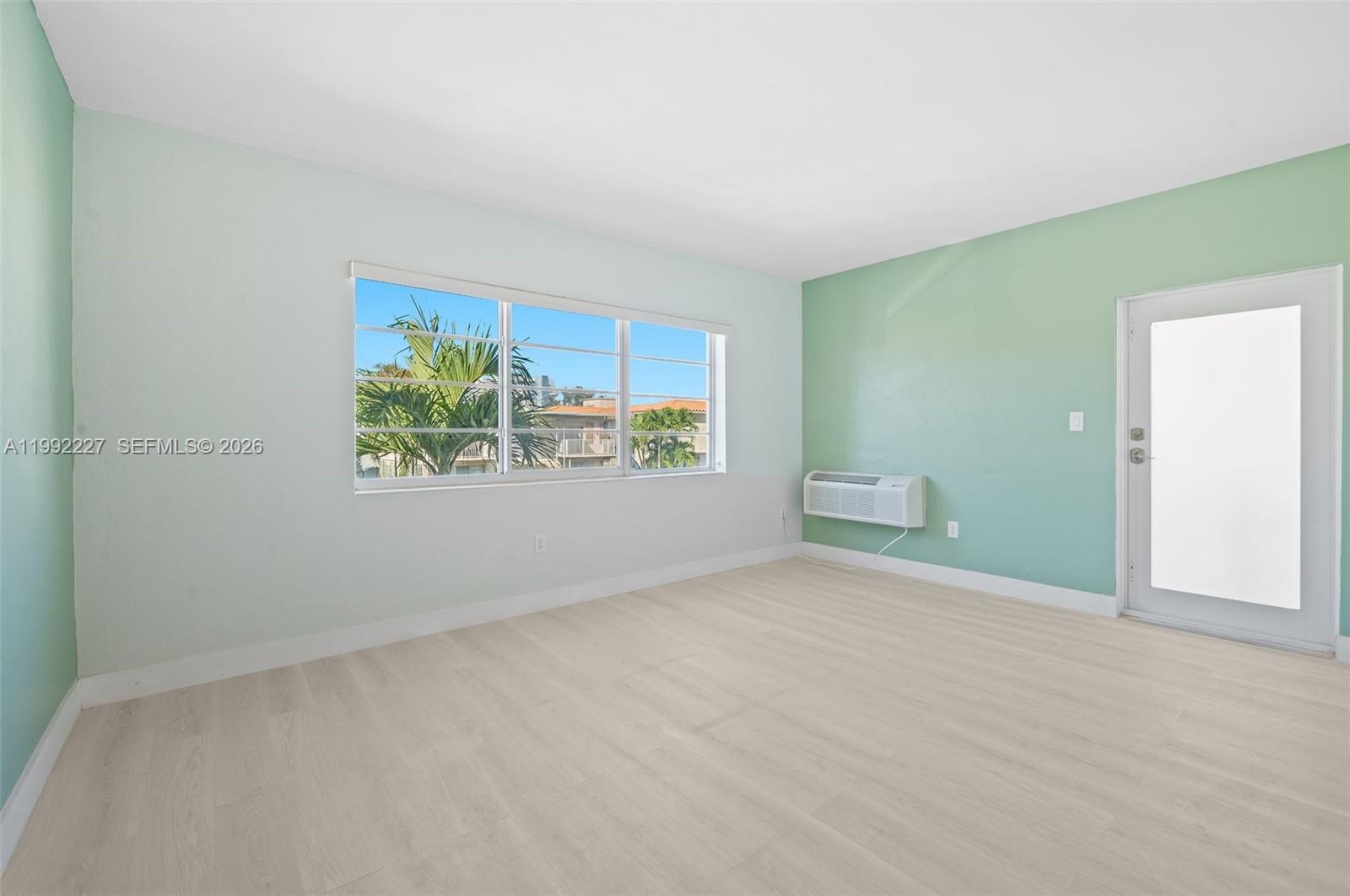 An empty room features light green walls, light wood-look flooring, a window, and an air conditioner — 7315 Byron Ave # 10, Miami Beach FL 33141, MIAMI BEACH, FL