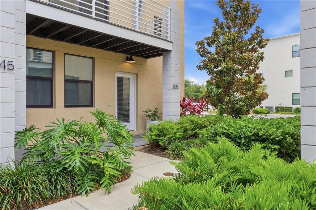 A beige townhouse entrance features a white door, a balcony above, and lush landscaping — 2551 11TH PL N, ST PETERSBURG, FL