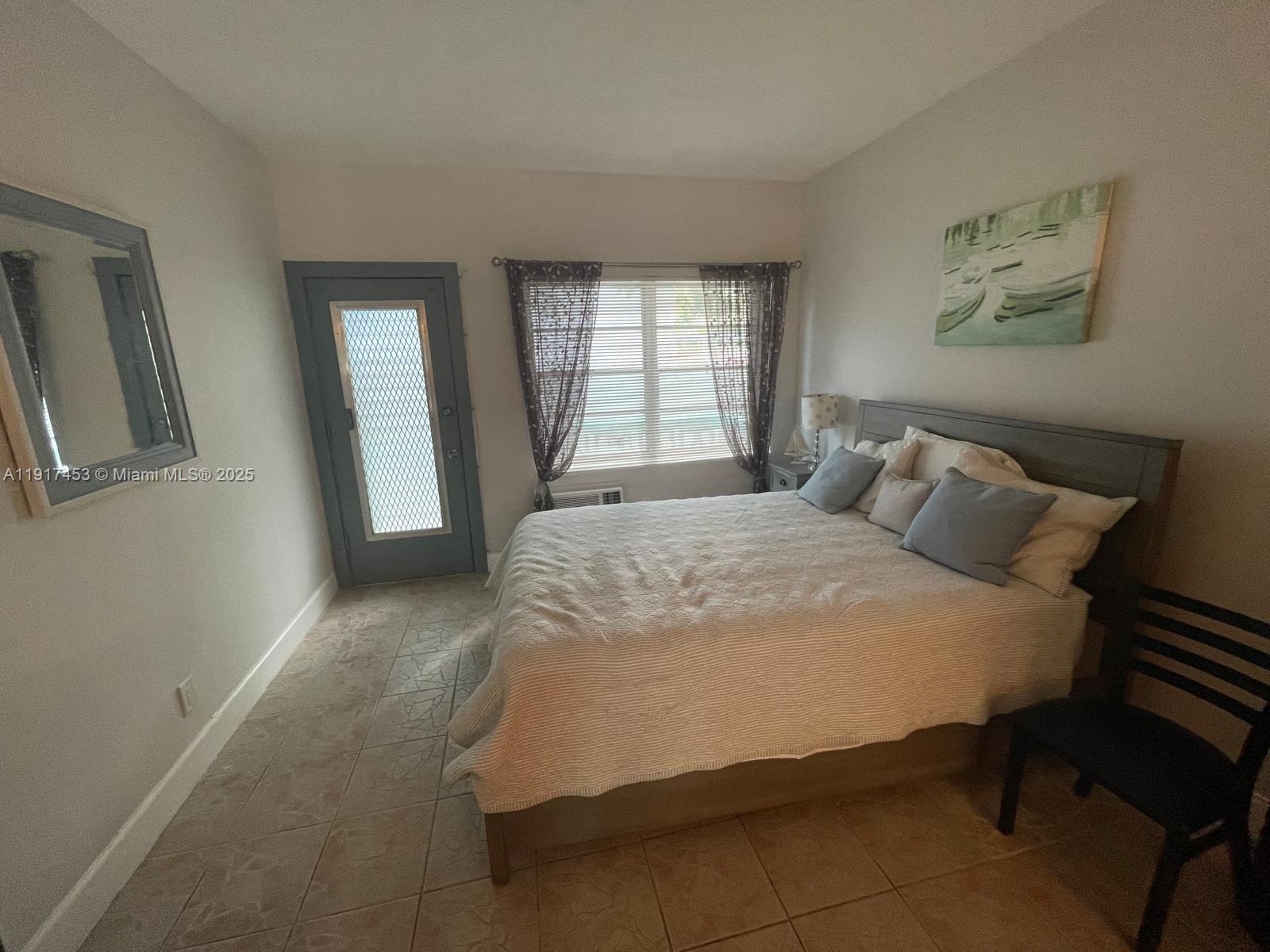 A bedroom features a bed, a blue door, a window with curtains, and tiled flooring — 6490 Collins Ave # 5, Miami Beach FL 33141, MIAMI BEACH, FL