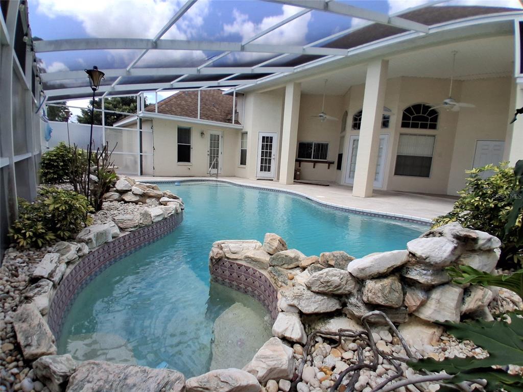 A screened-in swimming pool with a rock waterfall feature sits on a paved patio — 3392 ST IVES BOULEVARD, SPRING HILL, FL