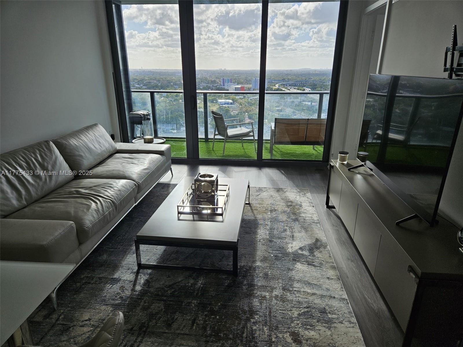 A modern living room features a leather sofa, coffee table, and balcony with city views — 121 NE 34 ST # 2511, Miami FL 33137, MIAMI, FL
