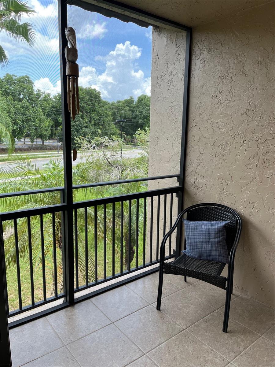 A screened-in balcony features tiled flooring, a black metal chair with a pillow, and greenery — WESTON, FL