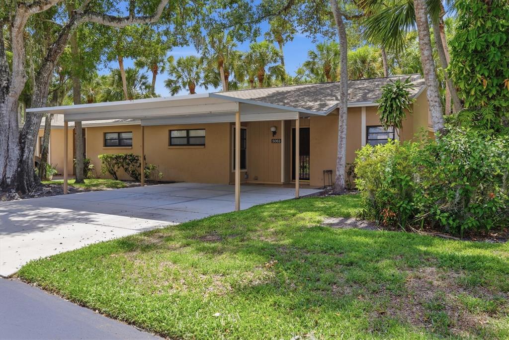 A single-story house with a tan exterior, a concrete carport, and surrounding trees and landscaping — 5063 VILLAGE GARDENS DRIVE UNIT 9, SARASOTA, FL