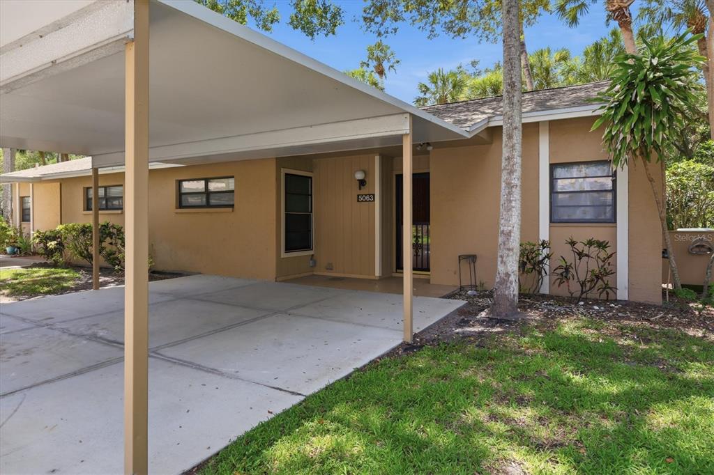 A tan single-story house features a concrete carport and a front entrance with a screen door — 5063 VILLAGE GARDENS DRIVE UNIT 9, SARASOTA, FL