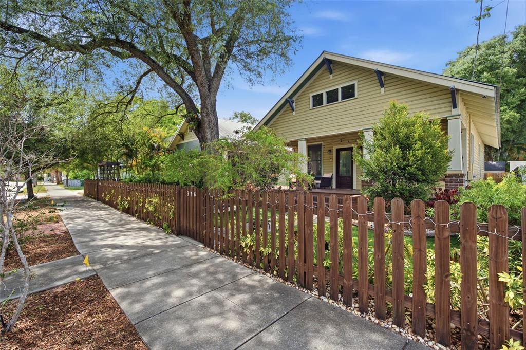 A yellow bungalow with a wooden picket fence sits behind a sidewalk and large tree — 316 E FRANCES AVENUE, TAMPA, FL