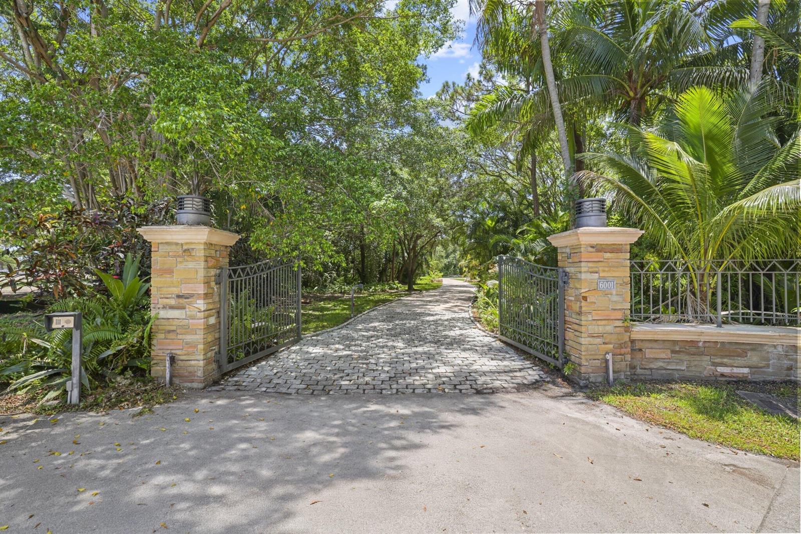 A stone-paved driveway leads through an open metal gate flanked by stone pillars and trees — 6001 Le Lac Road, Boca Raton FL 33496, BOCA RATON, FL