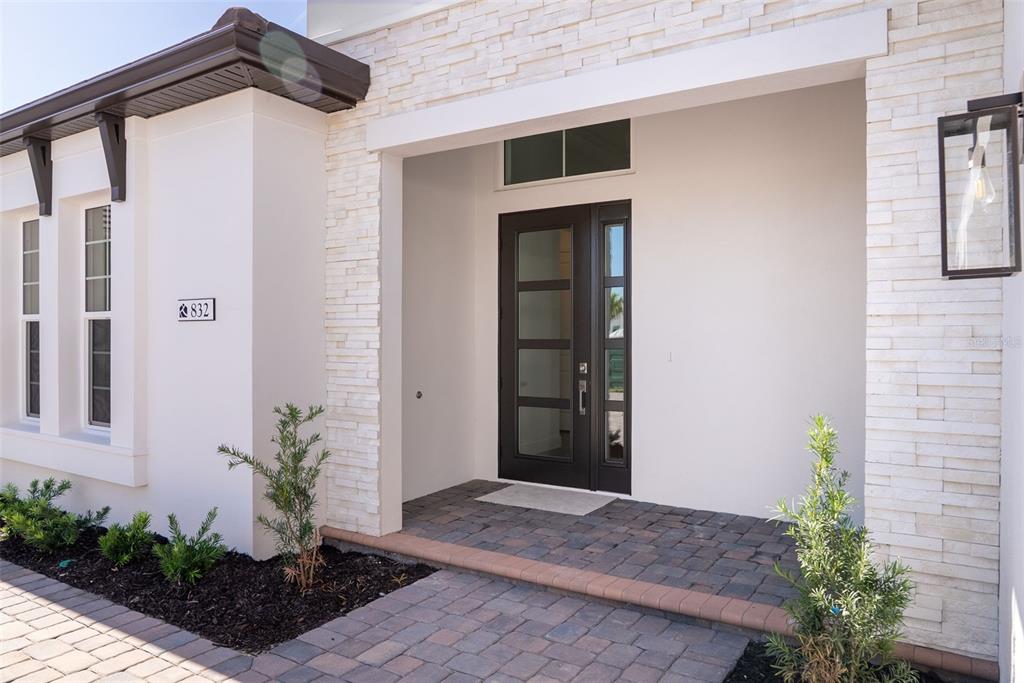 A modern home entrance features a dark front door, stone veneer accents, and paver walkway — 832 BLUE SHELL LOOP, SARASOTA, FL
