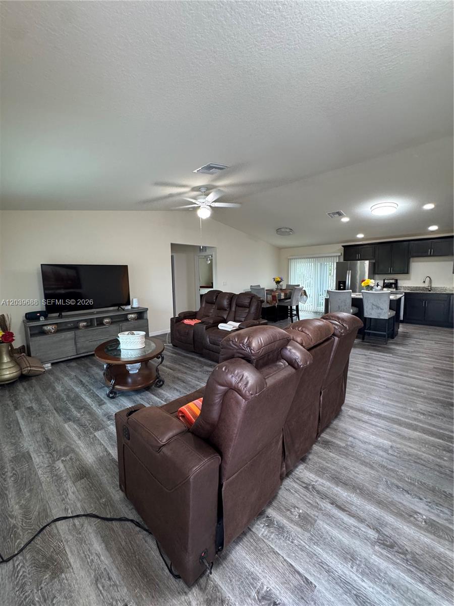 An open-concept living room features wood-look flooring, brown leather reclining furniture, and a kitchen area — 1201 BANJO CT, La Belle FL 33935, LA BELLE, FL
