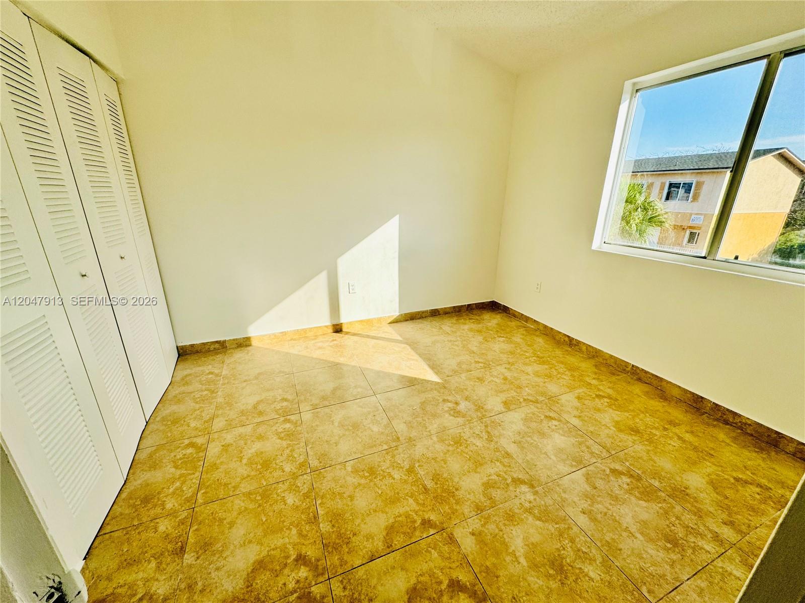 An empty bedroom features beige tile flooring, white walls, louvered closet doors, and a window — 6245 Kendale Lakes Cir # 208, Miami FL 33183, MIAMI, FL