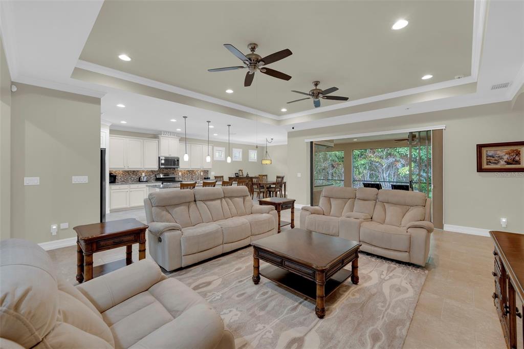 A spacious open-concept living room features beige furniture, a tray ceiling, and a kitchen beyond — 8720 WESTWOOD OAKS PLACE, FORT MYERS, FL