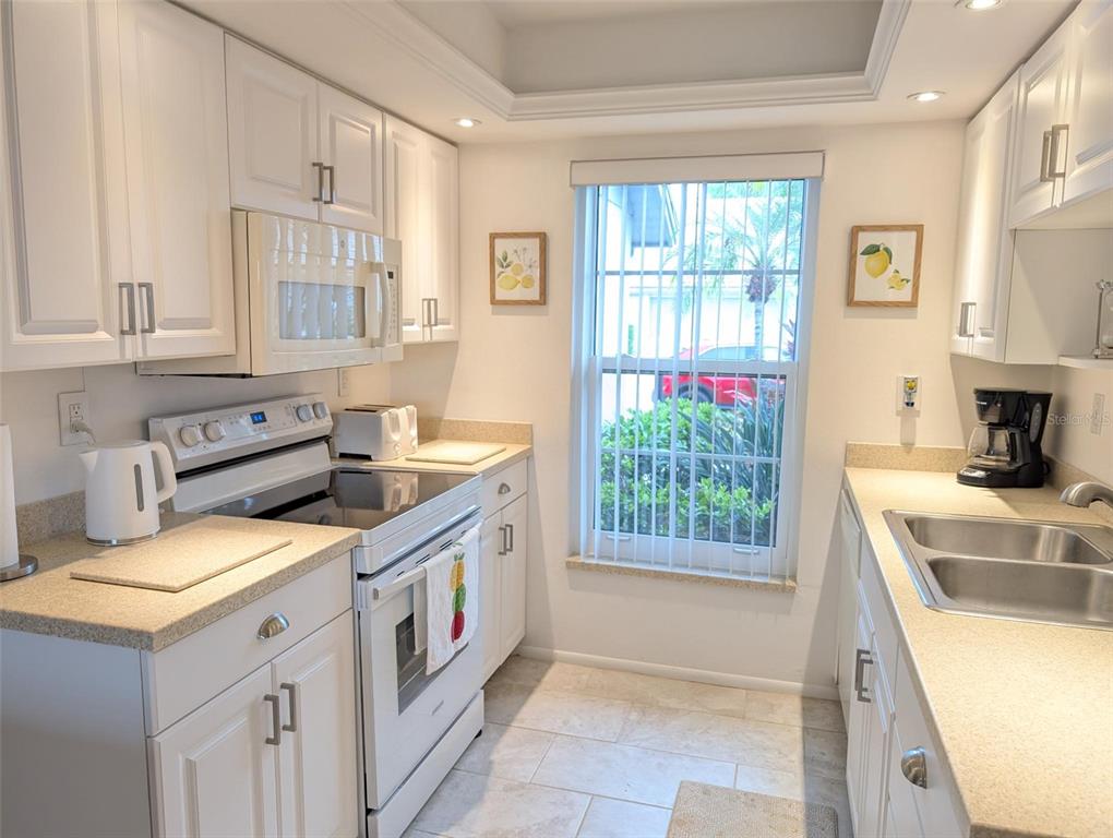 A white galley kitchen features light countertops, white cabinetry, and a window with vertical blinds — 5377 CRESTLAKE BOULEVARD UNIT 69, SARASOTA, FL