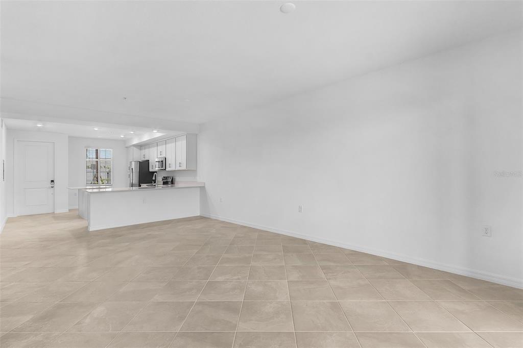 A spacious, empty room with light-colored tile flooring and an open-concept kitchen in the background — 14191 HERITAGE LANDING BOULEVARD UNIT 1214, PUNTA GORDA, FL