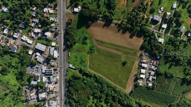 An aerial view shows a large, undeveloped grassy lot bordered by a road and houses — PR 2 KM. 120.6, AGUADILLA, PR