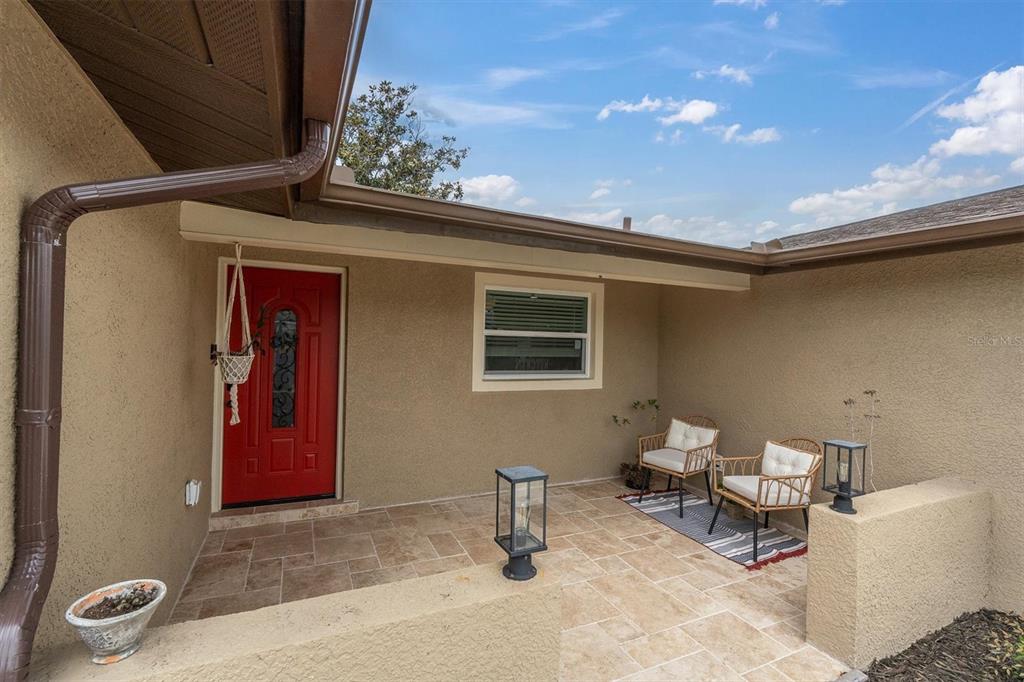 A covered patio features tiled flooring, two chairs on a rug, and a red front door — 8007 MIMOSA DRIVE, PORT RICHEY, FL