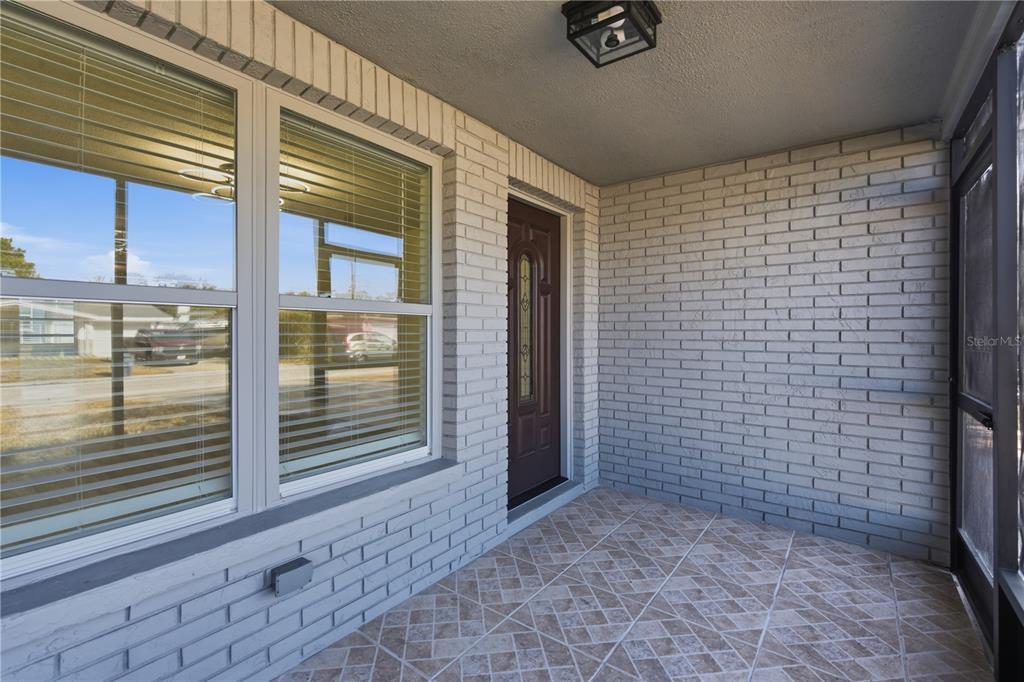 A covered porch with light-colored brick walls, tiled flooring, and a brown front door — 5330 POINSETTIA DR, NEW PORT RICHEY, FL