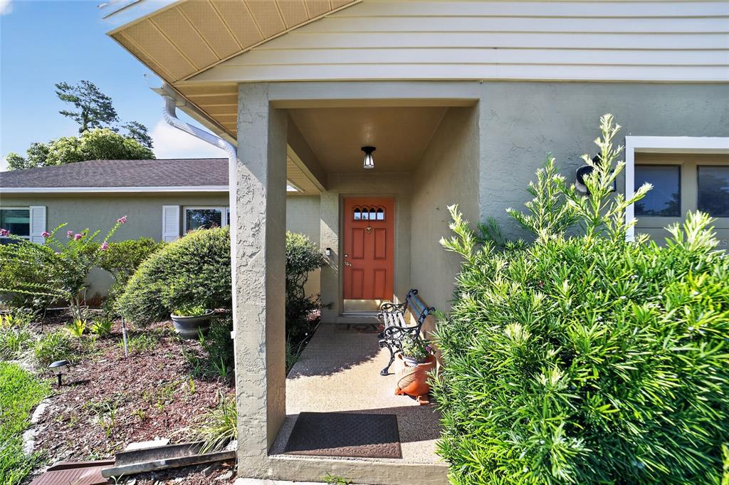 A covered entryway features a terracotta-colored front door, a small bench, and surrounding landscaping — 494 MARION OAKS MANOR, OCALA, FL