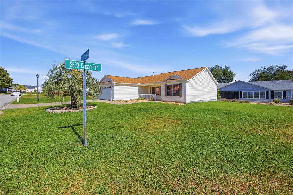 A single-story house with white siding and a tan roof sits on a grassy corner lot — 17705 SE 93RD HAWTHORNE AVENUE, THE VILLAGES, FL