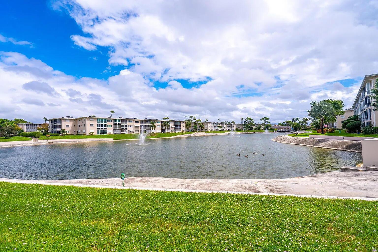 A pond with fountains sits in front of multi-story apartment buildings under a cloudy sky — 2606 Garden Drive S # 304, FL 33461, FL