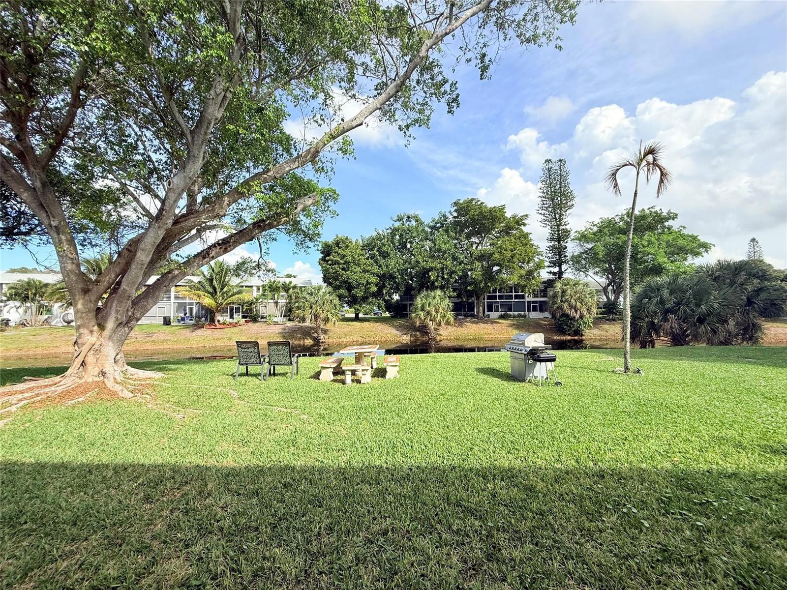 A grassy backyard features a large tree, a picnic table, chairs, and a grill by water — 53 Markham C # 53, Deerfield Beach FL 33442, DEERFIELD BEACH, FL