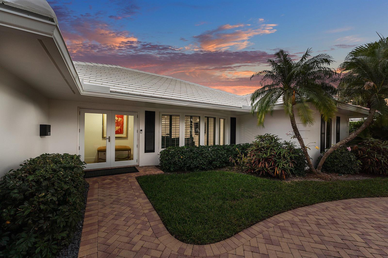 The exterior of a white house features a brick walkway, palm trees, and a sunset sky — 800 Holly Lane, Boca Raton FL 33486, BOCA RATON, FL