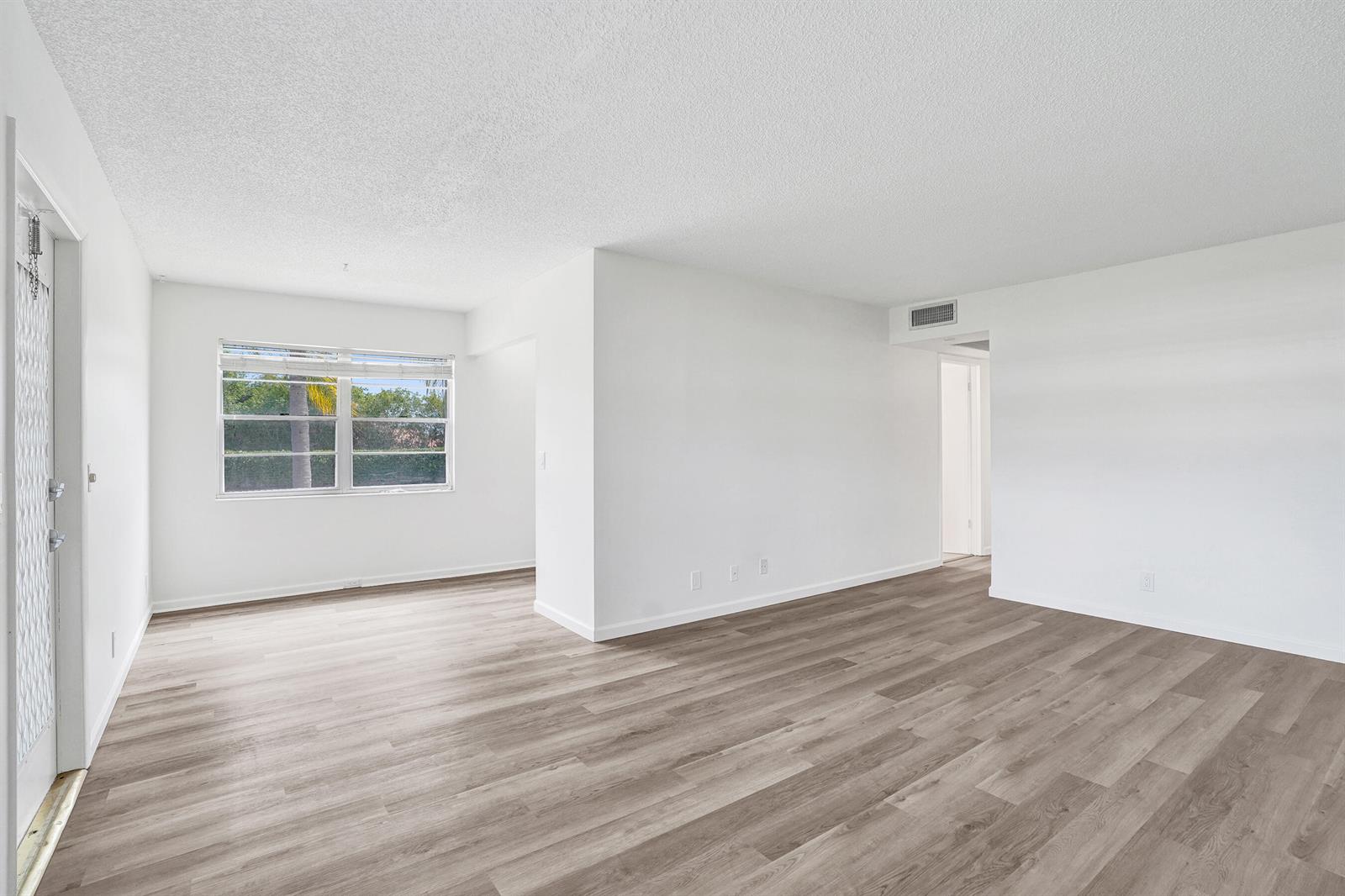 An empty living room features white walls, light wood-look flooring, and a window with blinds — 57 Dorset Drive # B, Boca Raton FL 33434, BOCA RATON, FL