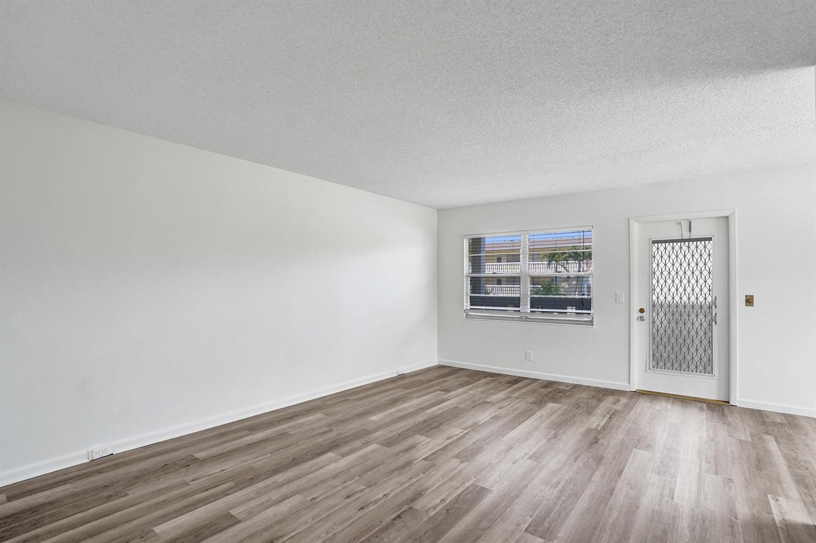 An empty room features white walls, light wood-look flooring, a window, and a glass-paneled door — 57 Dorset Drive # B, Boca Raton FL 33434, BOCA RATON, FL