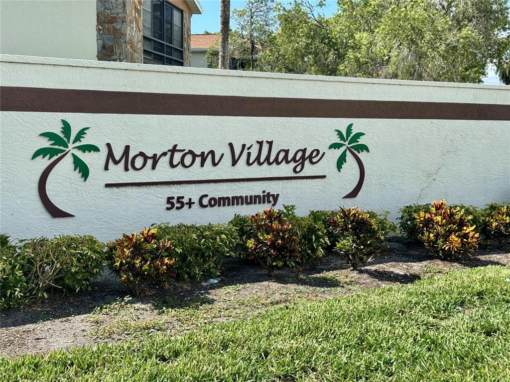 A white stucco wall features the Morton Village 55+ Community sign with palm tree accents — 5400 34TH STREET W UNIT 12H, BRADENTON, FL