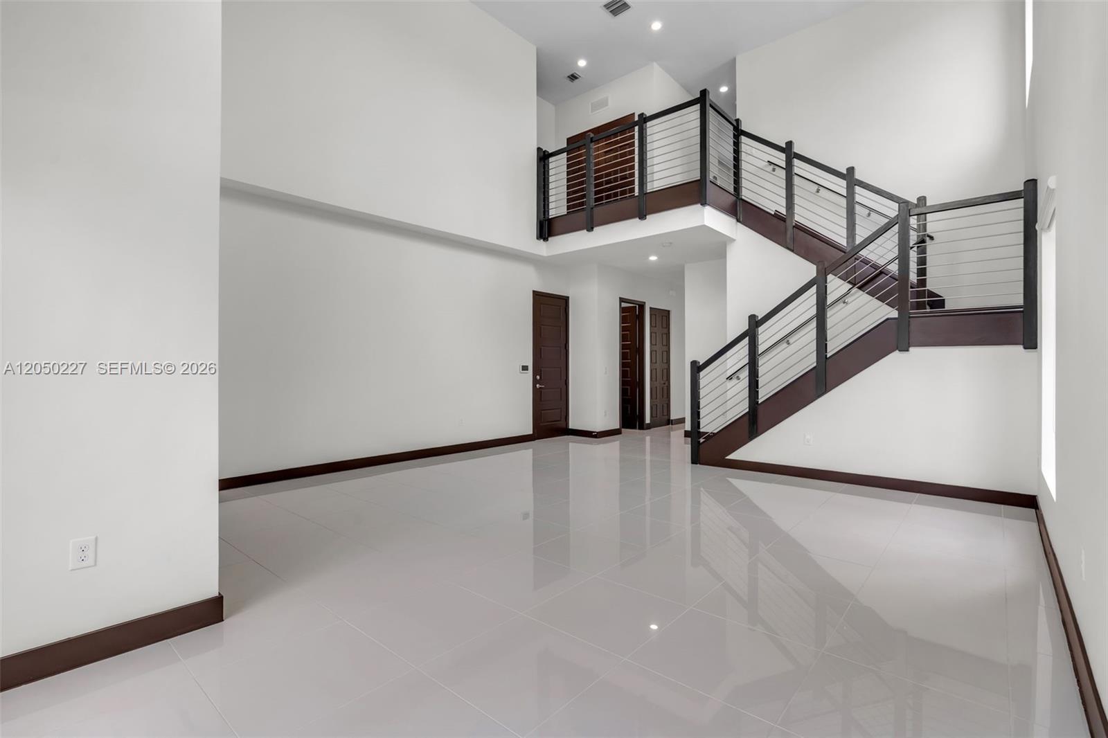A spacious, minimalist room features white tile floors, high ceilings, and a dark wood staircase — 15455 NW 88th Ct, Miami Lakes FL 33018, MIAMI LAKES, FL