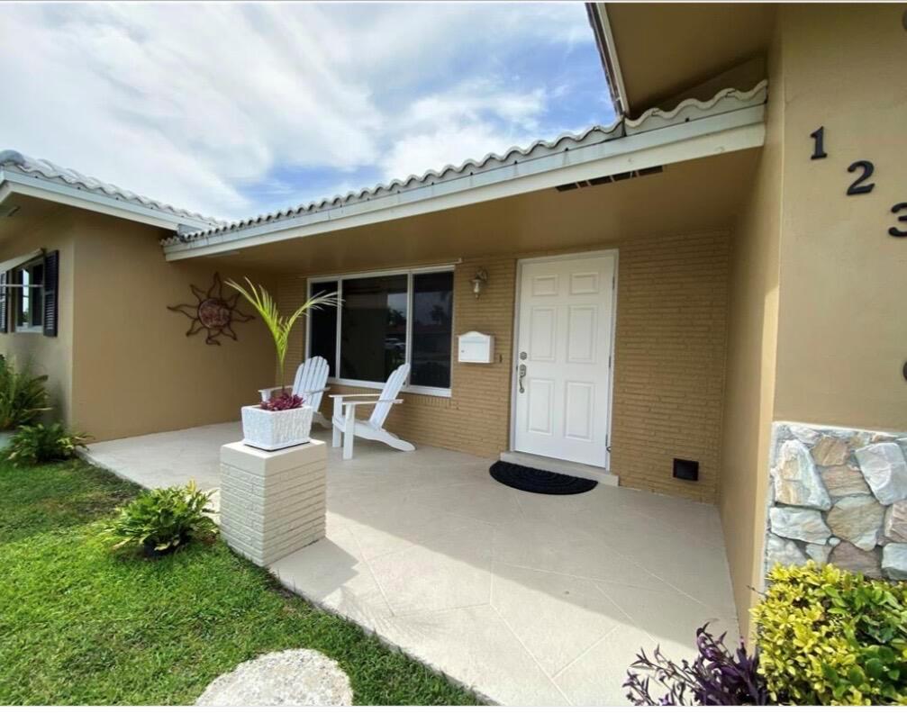 A tan house exterior features a covered front porch with white chairs and a door — 1230 SE 14th Drive, Deerfield Beach FL 33441, DEERFIELD BEACH, FL