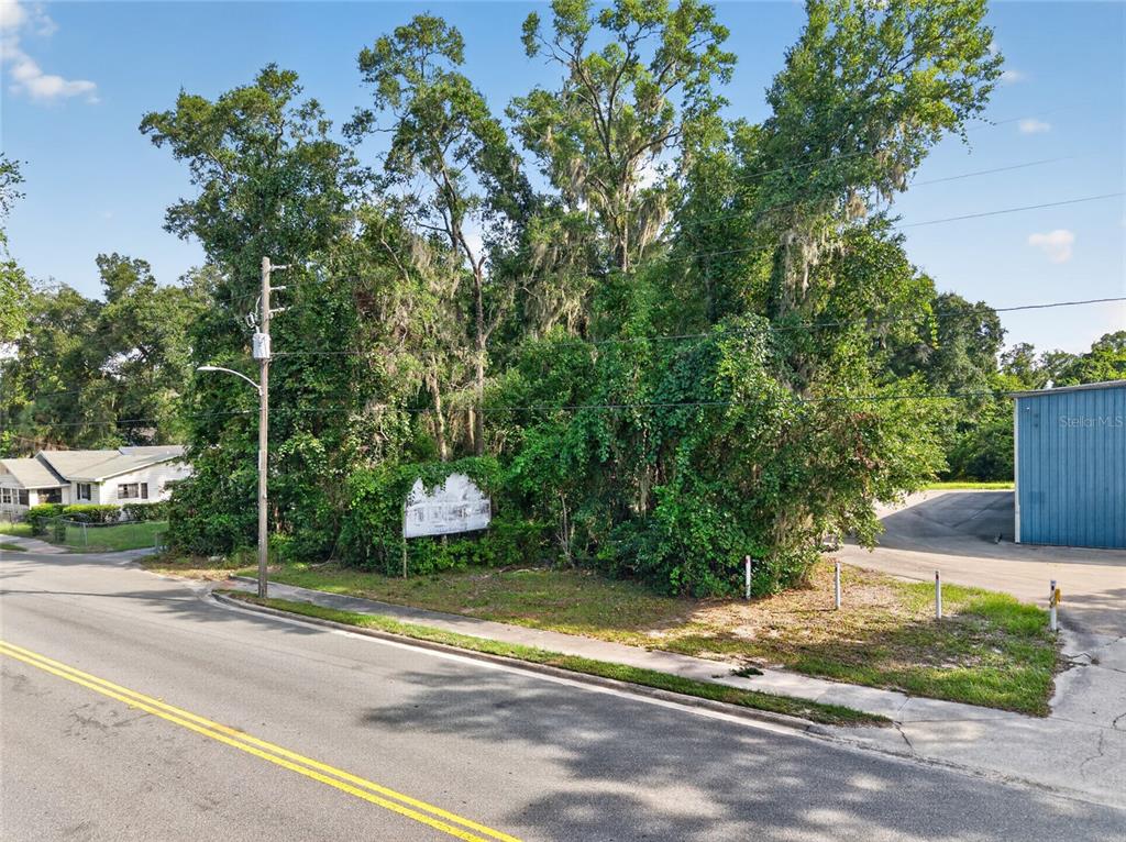 A vacant, tree-covered lot sits between a residential house and a blue industrial building — TBD NW 1ST AVENUE, HIGH SPRINGS, FL