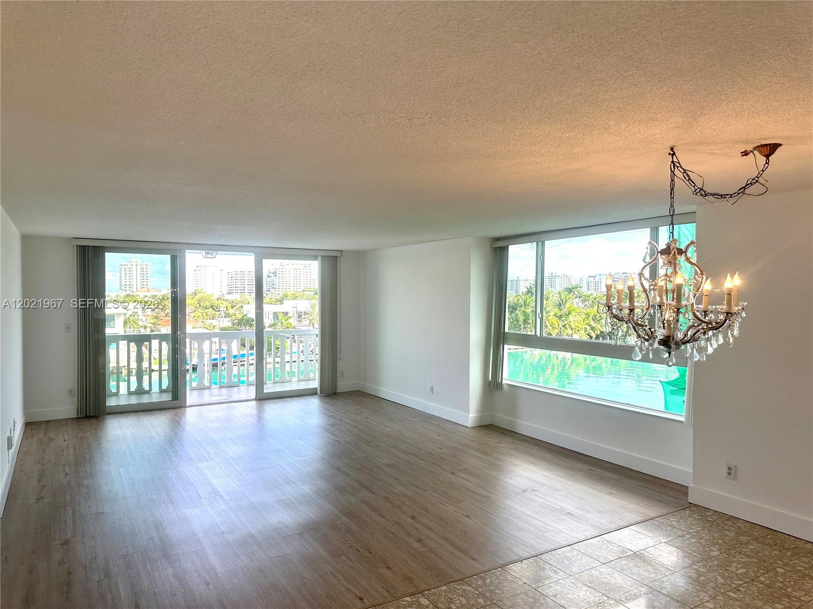An empty living room features wood-look flooring, a crystal chandelier, and large windows overlooking water — 9501 E Bay Harbor Dr # 5B, Bay Harbor Islands FL 33154, BAY HARBOR ISLANDS, FL