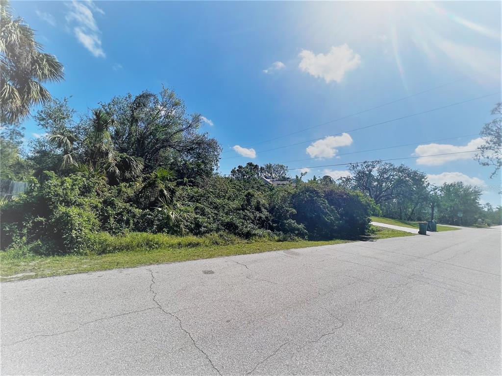 A vacant, overgrown residential lot sits along a paved street under a sunny sky — 3438 DUNKIRK STREET, PORT CHARLOTTE, FL